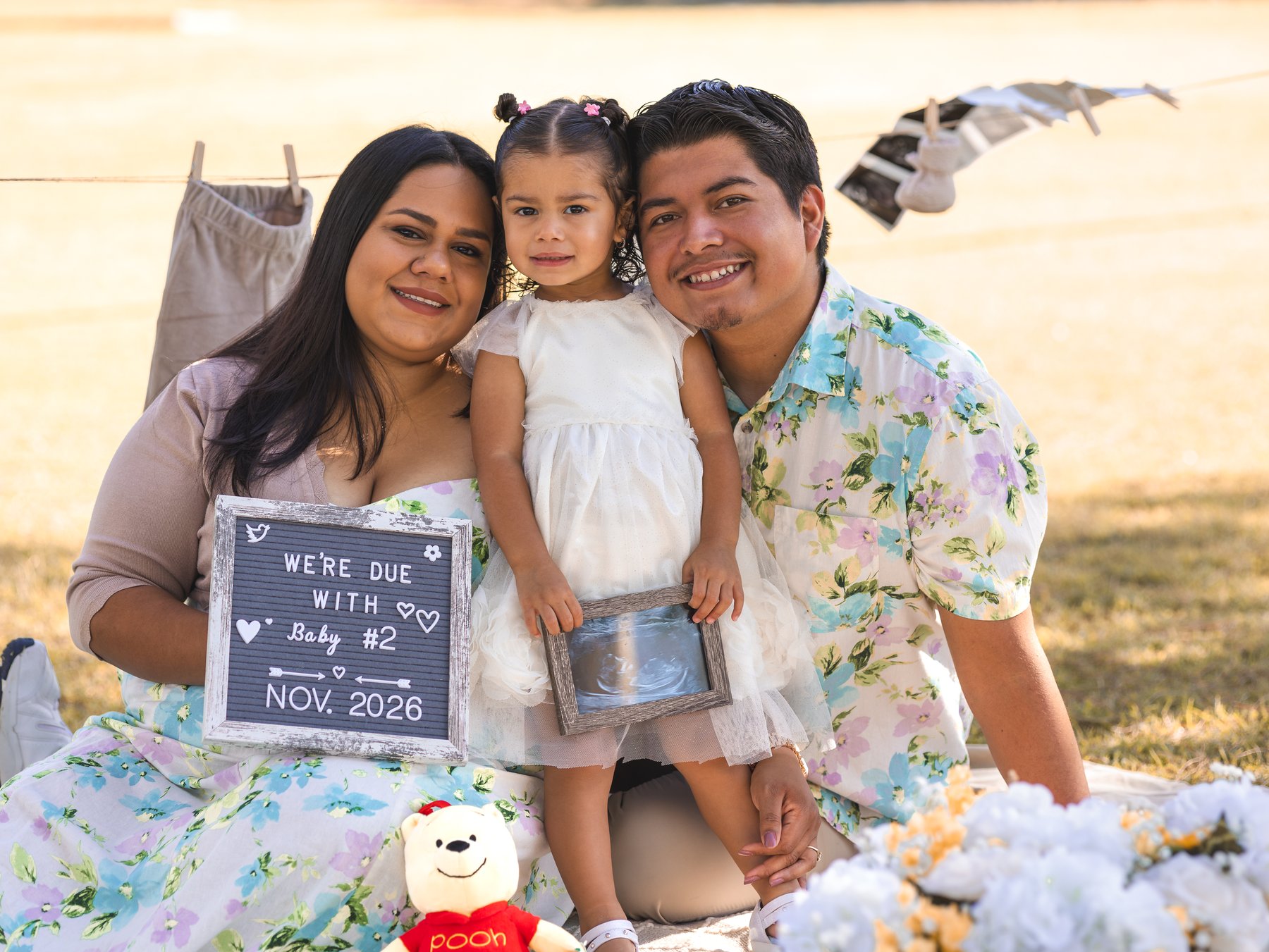 Mando and Mari with their child, an announcement sign and a framed sonogram outdoors